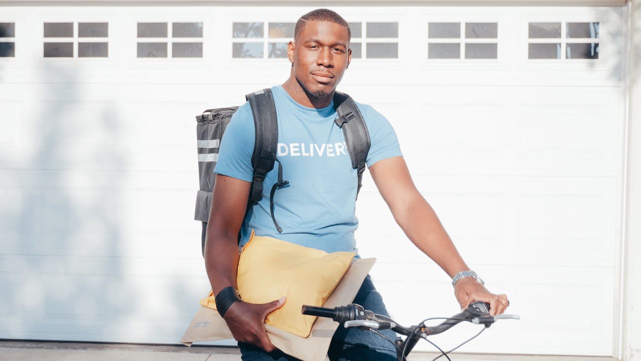 African American courier on bicycle holding parcels, ready for delivery in a sunny outdoor setting.